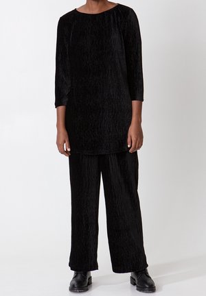 Persoon die een zwart getextureerd tuniek met lange mouwen en bijpassende wijde broekspijpen draagt, met zwarte chunky veterschoenen, op een witte achtergrond.