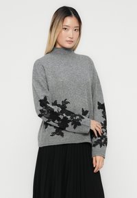 Femme portant un pull en tricot gris avec des broderies florales noires sur les manches et un col montant, associé à une jupe plissée noire.