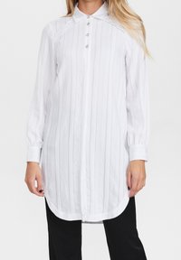 Vit långärmad skjorta med vertikala ränder, knappad krage, framknappar och rundad nederkant; tillverkad av lättviktsmaterial.