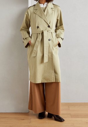 Trench beige con cinturón, llevado sobre camisa blanca y suéter beige, combinado con pantalones anchos marrones y botas negras de tacón.