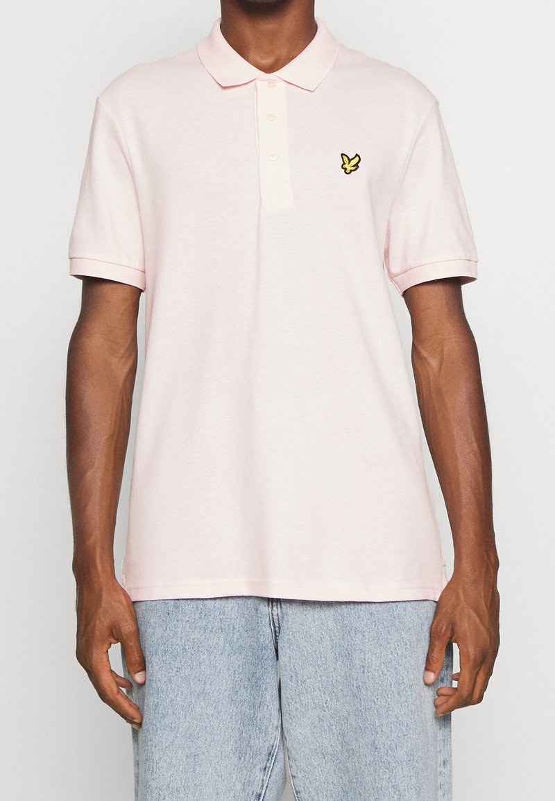 Homme portant un polo à manches courtes rose clair avec un logo d'oiseau jaune sur la poitrine et un jean en denim bleu clair, se tenant contre un fond uni.