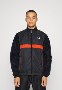 Chaqueta cortavientos negra con cremallera frontal, que presenta una franja horizontal naranja y un logo en el pecho, puños elásticos y dobladillo.
