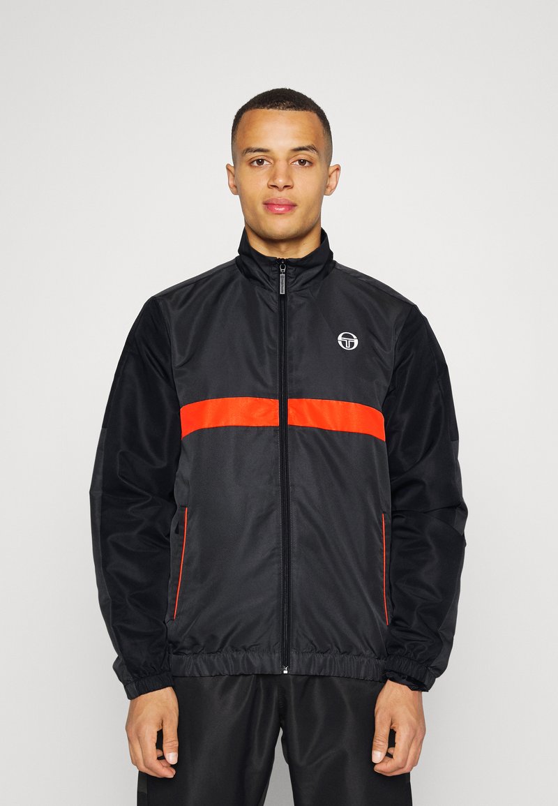 Chaqueta cortavientos negra con cremallera frontal, que presenta una franja horizontal naranja y un logo en el pecho, puños elásticos y dobladillo.