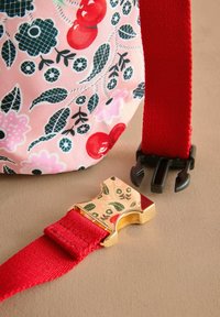 Stof met bloemenpatroon in roze, versierd met rode kersen en zwarte bloemen. Een gouden metalen gesp bevestigt een rode band met zwarte hardware.