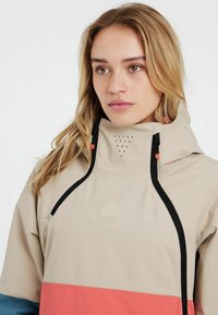 Helle beigefarbene Jacke mit einem korallenfarbenen unteren Bereich, ausgestattet mit einem hohen Kragen, einem schwarzen Reißverschluss und Belüftungsöffnungen, hergestellt aus glattem Stoff.