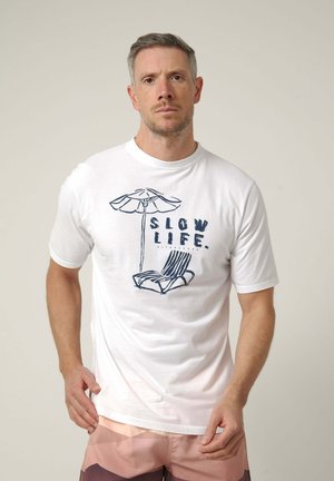 Hombre con cabello corto que lleva una camiseta blanca con un gráfico de paraguas y silla de playa y el texto "SLOW LIFE", combinado con pantalones cortos rosa claro.