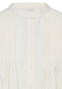 Blouse texturée écrue avec des plis verticaux, col rond et cinq boutons blancs au centre devant.