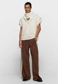 Mujer con un suéter de cuello alto blanco y peludo de manga corta y pantalones sueltos marrones con rayas laterales crema y negras, de pie con la mano en el bolsillo.