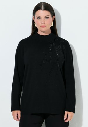 Vrouw met steil donker haar draagt een zwarte trui met lange mouwen met een subtiel kralenpatroon en gouden oorbellen, staand tegen een witte muur.