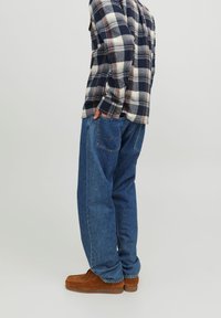 Camicia di flanella a quadri in navy, bianco e ruggine; jeans blu larghi; scarpe in suede marroni; mani nelle tasche; vista laterale.
