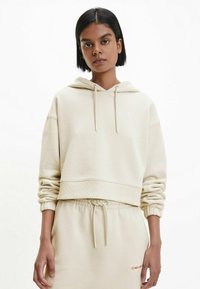 Sudadera cortada beige en tejido suave con capucha ajustable. Presenta puños y dobladillo acanalados, con un pequeño logo bordado en el pecho.