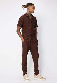 Ensemble de chemise à manches courtes et de pantalon marron fabriqué en tissu texturé. Dispose d'un col et d'une fermeture par bouton. Associé à des baskets beige.