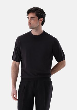 Mann mit dunklen Haaren, der ein schwarzes Kurzarmshirt mit Rundhalsausschnitt und schwarze Hose trägt, schaut nach links vor neutralem Hintergrund.