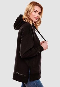Sudadera negra con cordón y aberturas laterales, con una textura suave y detalle bordado en el dobladillo. El modelo lleva puestos unos jeans azules.