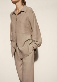 Beige button-up shirt met een kraag, één borstzak en een relaxte pasvorm, gecombineerd met bijpassende taps toelopende broek. Een gladde textuur, minimalistisch ontwerp.