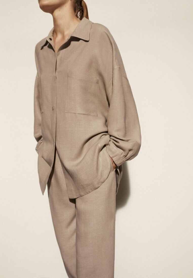 Beige button-up shirt met een kraag, één borstzak en een relaxte pasvorm, gecombineerd met bijpassende taps toelopende broek. Een gladde textuur, minimalistisch ontwerp.