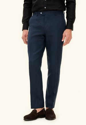 Man wearing navy blue tailored trousers, black shirt tucked in, and dark brown suede loafers, standing against plain background.