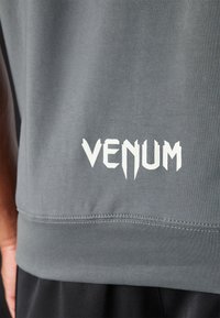 Sivá atletická tričká s bielym logom "VENUM" na leme. Tkanina je hladká s priliehavým dizajnom a krátkymi rukávmi.