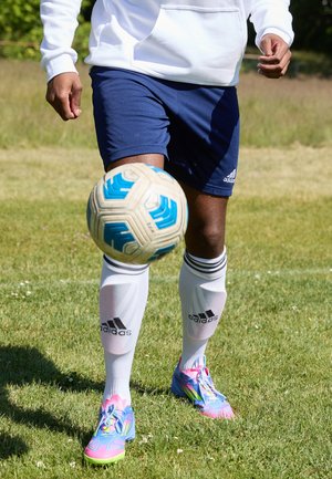 Personne portant un short bleu, des chaussettes blanches Adidas et des crampons colorés jonglant avec un ballon de football blanc et bleu sur l’herbe.