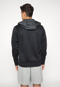 Svart huvtröja med en texturerad huva, mjukt tyg, åtsittande ärmmuddar och en snygg design. Får bäras med grå shorts, vilket visar bakvyn.