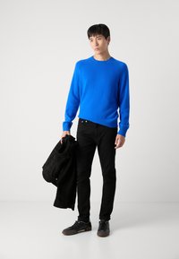Suéter azul de punto con puños acanalados, combinado con jeans ajustados negros y zapatillas negras, sosteniendo una chaqueta negra texturizada.