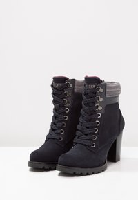 Námořnicky modré semišové kotníkové boty s robustním podpatkem, šněrováním vpředu, stříbrnými průchodkami a texturovaným lemováním. Gumová podrážka pro lepší trakci a odolnost.