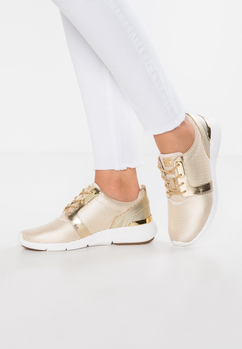 Goldene Mesh-Sneaker mit metallischen Akzenten, Schnürdesign, weißer Sohle und strukturierten Paneelen, kombiniert mit weißen, ausgefransten Jeans.