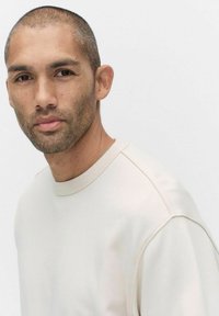 Jasno beżowy sweatshirt z fakturowanym, okrągłym dekoltem. Materiał wydaje się miękki, z subtelnymi detalami wszycia i luźnym krojem.