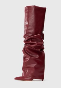 Bota de cuero burdeos alta y holgada con punta afilada y tacón bajo sobre un fondo ligero liso.