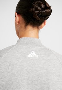 Grå sweatshirt med høj krave, lavet af et tekstureret stof. Bemærkelsesværdigt hvidt Adidas-logo på ryggen nær halsudskæringen.