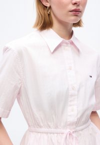 Γυναίκα που φοράει ανοιχτό ροζ φόρεμα με κοντά μανίκια και κουμπιά, με κορδόνι στη μέση και χρυσό σκουλαρίκι-κρίκο, φανερώνεται από τους ώμους μέχρι τη μέση.