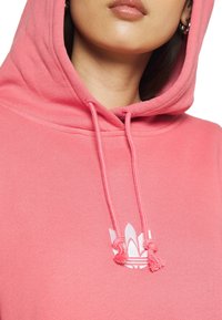 Růžová mikina s kapucí, vybavená šňůrkou s třásněmi a bílým potiskem loga. Materiál má jemnou texturu a volný střih.