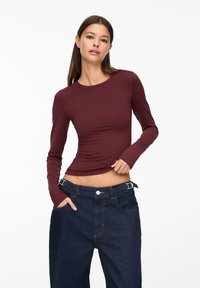 Bordeauxfarbene Langarm-Bluse aus glattem Stoff, körpernah geschnitten, kombiniert mit lockeren dunklen Jeanshosen mit einem breiten Bund.