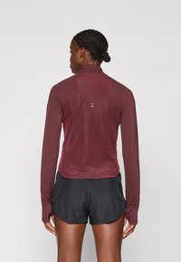 Bordeaux rotes langärmliges Sportoberteil mit strukturiertem Rücken, kombiniert mit schwarzen Shorts. Verfügt über einen Stehkragen und Daumenlöcher an den Ärmeln.