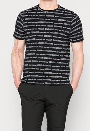 Homme portant un t-shirt noir Armani Exchange avec un logo blanc répétitif et un pantalon noir, les mains dans les poches, debout devant un fond blanc.