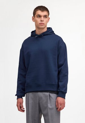 Mężczyzna o krótkich brązowych włosach ubrany w granatową bluzę z kapturem i szare plisowane spodnie, stojący na jednolitym białym tle.