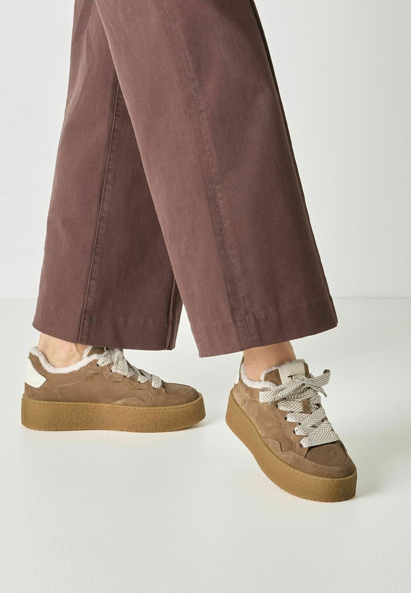 Personne portant un pantalon marron à jambes larges et des baskets compensées beige avec une doublure en fourrure blanche et des lacets épais à motifs sur un fond uni.