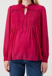 Blouse rose avec un col à volants, une ouverture en forme de goutte et des détails plissés. Manches longues élastiquées avec des accents texturés. Coupe fluide.