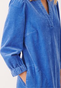 Abito chemisier in velluto a coste blu con scollatura a V, delicati righe verticali, maniche lunghe con polsini arricciati e tasche laterali. Morbido al tatto.