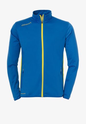 Blå jakke med høj krave, fuldlængde gul lynlås, sidelommer og brændmærke i gult på venstre bryst. Glat tekstur.