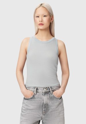 Jonge vrouw met lang blond haar, gekleed in een lichtgrijze mouwloze geribbelde top en vervaagde grijze spijkerbroek met handen in de zakken.