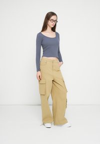 Bluza albastră din bumbac, cu mâneci lungi și croială mulată, asortată cu pantaloni largi bej cu buzunare laterale. Modelul poartă adidași albi.