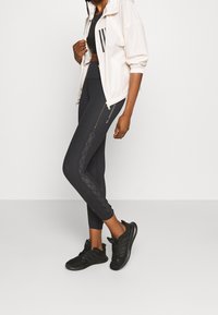 Svarta sporttights med texturerade sidopaneler, i kombination med en ljusbeige zip-up jacka. Svarta sneakers fulländar outfiten.