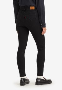 Personne portant un jean skinny noir, une chemise noire, des chaussettes blanches et des mocassins noirs, vue de dos sur un fond blanc.