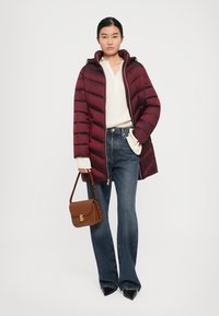 Vrouw in een bordeauxrode pufferjas, witte trui, blauwe spijkerbroek, zwarte puntige schoenen, met een bruine handtas met een gouden sluiting.