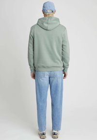 Un sweat à capuche vert avec une poche kangourou et un cordon de serrage, associé à un jean bleu clair et des baskets noir et blanc. Vue de dos.