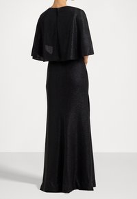 Svart aftonklänning med ett flödande, kappa-liknande överdrag. Har en elegant silhuett, texturerad tyg och en dragkedja bak.