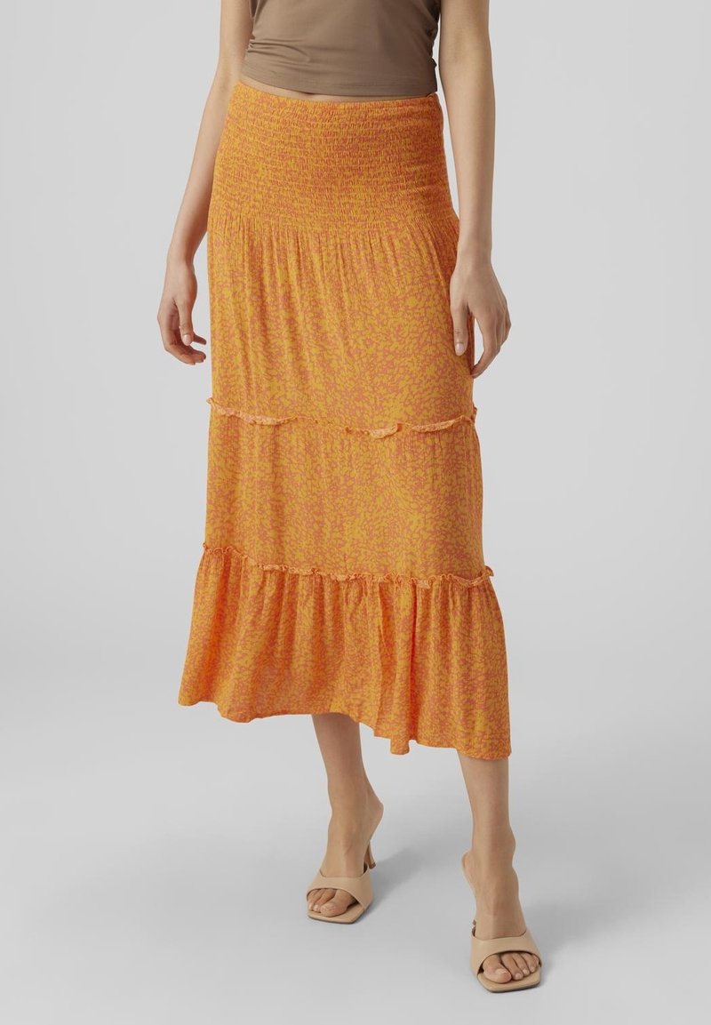 Falda maxi de color naranja con estampado, con cintura fruncida, que presenta volantes en capas y una tela ligera y texturizada. Combinada con sandalias abiertas en color beige.
