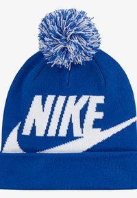 Kék kötött sapka fehér „NIKE” logóval. Texturált felülettel és nagy, bolyhos fehér-kék pomponnal a tetején.
