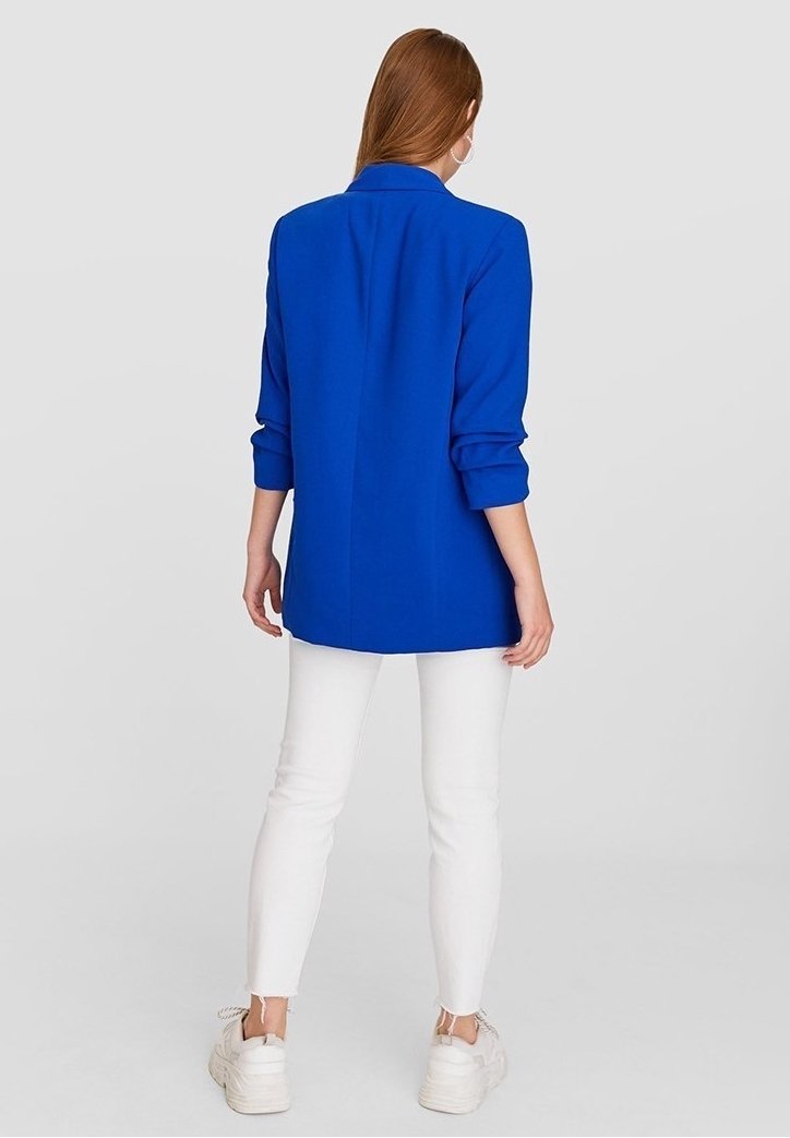 Blå kavaj med krage, uppvikta ärmar och rak passform, bärs över vita byxor och kraftiga vita sneakers. Slät tygtextur.